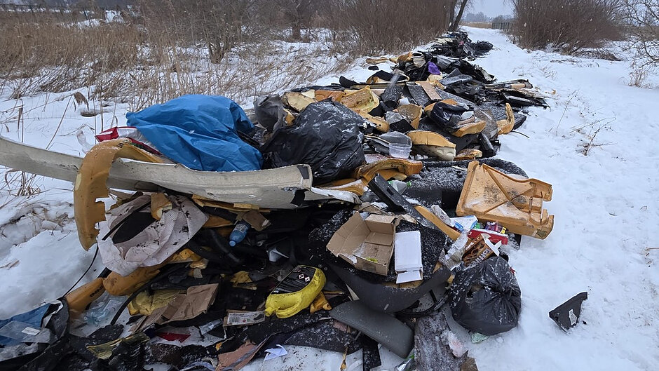 Na zdjęciu widać dużą stertę porzuconych śmieci zalegającą przy ośnieżonej, wiejskiej drodze. Wśród odpadów znajdują się czarne worki na śmieci, kawałki pianki i tapicerki, kartony, plastikowe pojemniki oraz inne różnorodne elementy. Całość leży na tle zimowego krajobrazu – wokół widać śnieg, suche trawy i bezlistne drzewa. Scena sprawia wrażenie nielegalnego wysypiska, które wyraźnie kontrastuje z naturalnym otoczeniem.