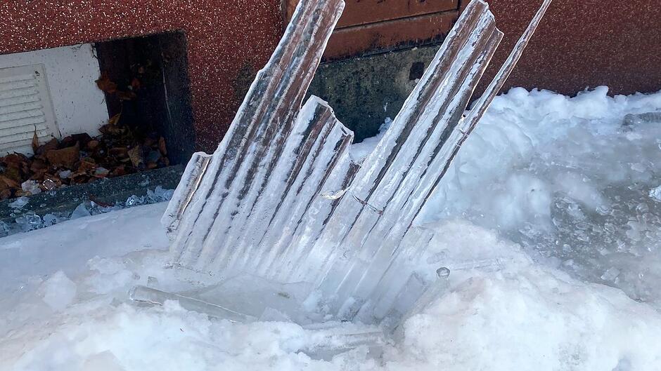 Na zdjęciu widać duże, ostre fragmenty lodu wbite w śnieg tuż przy ścianie budynku. Lodowe elementy mają postrzępione krawędzie i wyglądają jak pozostałości po dużym soplu lub pękniętej bryle, która spadła z wysokości. W tle widoczny jest fragment elewacji i zamarznięta warstwa śniegu, podkreślające skalę zagrożenia stwarzanego przez spadające sople.