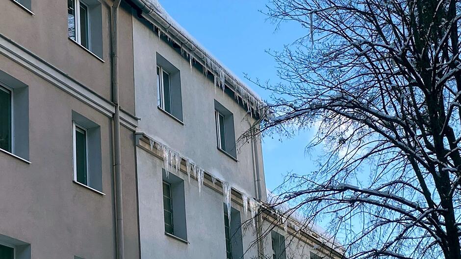 Na zdjęciu widać fragment elewacji budynku mieszkalnego, z którego krawędzi dachu oraz gzymsów zwisają długie sople. Lodowe zwisy tworzą się na całej długości ściany, a część z nich osiąga znaczną długość. Po prawej stronie kadru widoczne są gałęzie drzewa pokryte cienką warstwą lodu. Jasne niebo i słońce podkreślają kontrast między elewacją a ostrymi, niebezpiecznymi soplami.