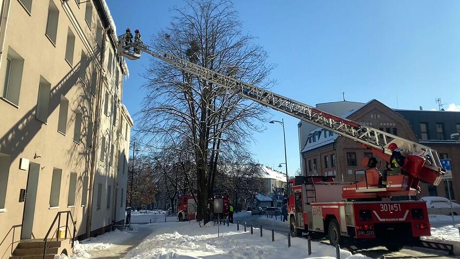 Na zdjęciu widać straż pożarną podczas usuwania sopli z dachu budynku mieszkalnego. Strażacy znajdują się na podnośniku umieszczonym na wysuniętej, wysokiej drabinie zamontowanej na wozie strażackim. Pojazd stoi na ośnieżonej ulicy, a w tle widać inne wozy strażackie oraz zabudowę miejską. Jasne, słoneczne niebo i zalegający śnieg podkreślają zimowe warunki, w których prowadzona jest akcja.