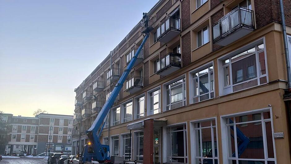 Na zdjęciu widać pracowników korzystających z podnośnika koszowego, którzy usuwają sople z krawędzi balkonów na fasadzie wielopiętrowego budynku. Podnośnik w kolorze niebieskim jest rozstawiony na ulicy, a jego wysięgnik sięga górnych kondygnacji. Wokół panują zimowe warunki — na chodnikach leży śnieg, a w tle widoczne są inne budynki mieszkalne. Scena pokazuje działania mające na celu zapewnienie bezpieczeństwa przechodniom.