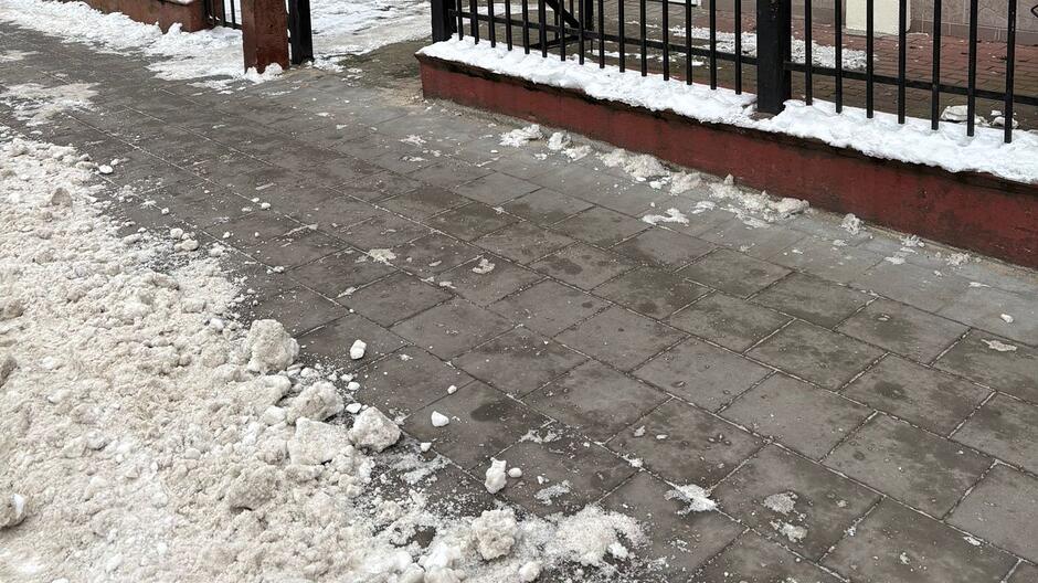 Na zdjęciu widać odśnieżony chodnik wzdłuż ogrodzenia. Płyty chodnikowe są w większości czyste, a śnieg został zgarnięty na krawędź po lewej stronie, gdzie utworzyła się niewielka pryzma zbitego, białego i lekko zabrudzonego śniegu. Przy krawężniku i przy murku pod ogrodzeniem leżą pojedyncze grudki śniegu, ale cała powierzchnia chodnika wygląda na starannie uprzątniętą i nadającą się do swobodnego przejścia. Całość sprawia wrażenie uporządkowanej i zadbanej przestrzeni w zimowych warunkach.