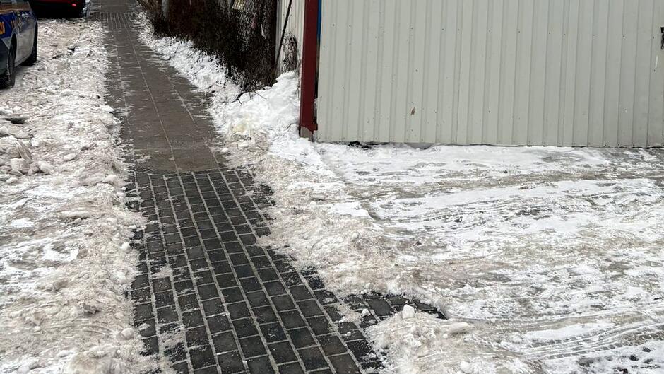 Na zdjęciu widać zimową scenę przedstawiającą odśnieżony chodnik biegnący wzdłuż ogrodzenia lub budynku. Ścieżka z ciemnej kostki brukowej została starannie oczyszczona i jest wyraźnie odróżniona od otaczającego ją rozjeżdżonego, zbitego śniegu. Po lewej stronie chodnika widoczne są resztki odgarniętego śniegu, a dalej widać fragment zaparkowanego samochodu.
Po prawej stronie znajduje się duża, jasna ściana (prawdopodobnie garażu lub budynku gospodarczego), przed którą zalega warstwa rozjeżdżonego, brudnego śniegu z widocznymi śladami opon. Ogółem zdjęcie pokazuje różnicę między dokładnie odśnieżoną częścią ciągu pieszego a nieuprzątniętym terenem obok.