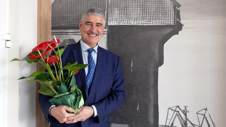 Na zdjęciu znajduje się osoba ubrana w elegancki, granatowy garnitur, niebieską koszulę oraz krawat w odcieniach niebieskiego. Trzyma w rękach bukiet roślin doniczkowych z czerwonymi kwiatami (prawdopodobnie anturium), owinięty w zielony papier dekoracyjny.
W tle widać dużą, czarno‑białą grafikę lub fotografię przedstawiającą elementy architektury przemysłowej — konstrukcję przypominającą budynek portowy lub magazyn oraz fragmenty dźwigów. Po lewej stronie kadru widać fragment ściany i drewnianego wykończenia. Całość sprawia wrażenie ujęcia wykonanego z okazji uroczystości, podziękowania lub wręczenia symbolicznego upominku.