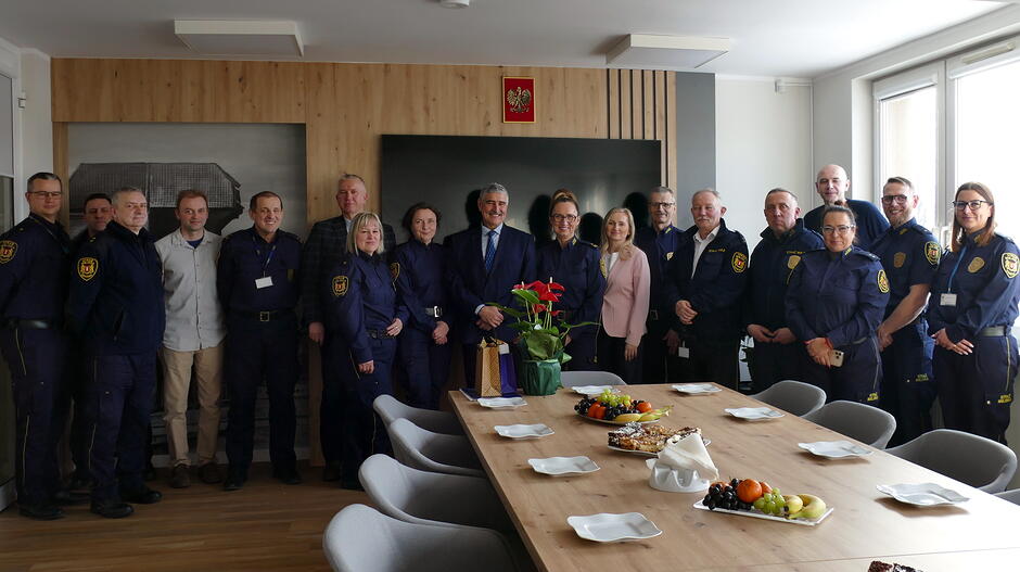 Na zdjęciu widać grupę kilkunastu osób stojących w pomieszczeniu o charakterze biurowo‑konferencyjnym. Większość z nich ma na sobie granatowe uniformy służbowe z emblematami na ramionach, a część trzyma identyfikatory na smyczach. W centrum grupy stoi osoba w eleganckim garniturze, obok której znajdują się również dwie osoby w stroju cywilnym.
Przed grupą ustawiony jest duży stół konferencyjny nakryty talerzami i półmiskami z owocami, słodkimi przekąskami i drobnymi poczęstunkami. Na środku stołu znajdują się również kwiaty oraz kilka upominkowych toreb. Pomieszczenie jest jasne, z dużymi oknami po prawej stronie. Na ścianie za grupą wisi telewizor oraz tablica z godłem.
Całość sprawia wrażenie spotkania służbowego lub uroczystego zebrania, być może z okazji święta, podziękowań lub oficjalnego wyróżnienia.