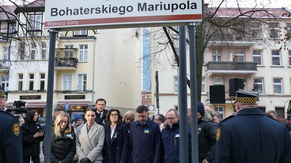 Na zdjęciu widać uroczystość odbywającą się na ulicy, przy tablicy z nazwą Bohaterskiego Mariupola w dzielnicy Wrzeszcz Górny. Pod znakiem stoi grupa osób, a wokół nich zgromadziło się więcej uczestników wydarzenia. W tle widoczne są kamienice oraz elementy miejskiej zabudowy. Obecni są również funkcjonariusze straży miejskiej w granatowych płaszczach.