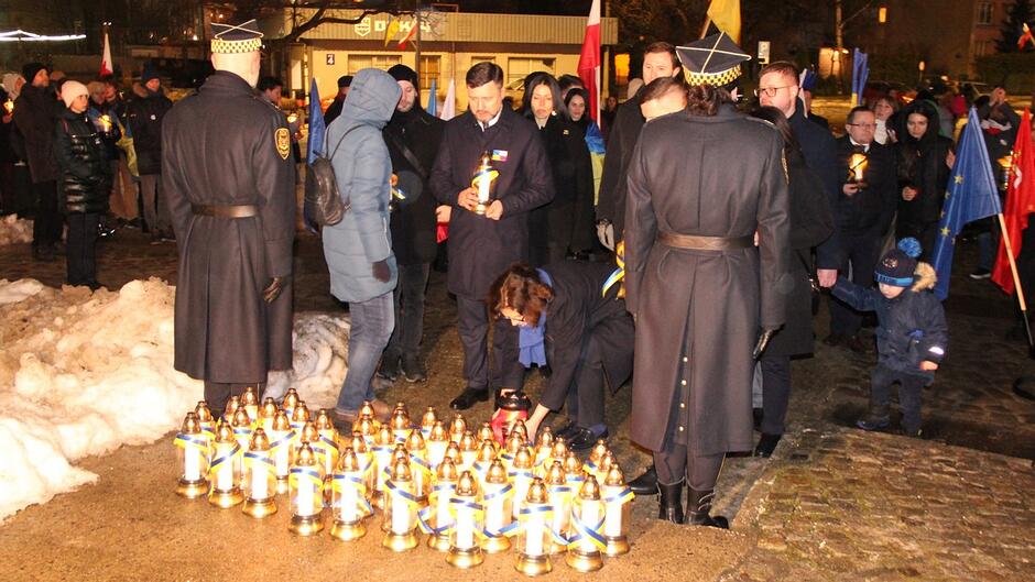 Zdjęcie przedstawia zgromadzenie w godzinach wieczornych lub po zmroku. Widać grupę osób stojących na placu, a na pierwszym planie liczne znicze ustawione w rzędach. Niektóre osoby trzymają znicze w dłoniach, a jedna osoba pochyla się nad ustawionymi lampionami, jakby je porządkowała lub zapalała. Wokół znajdują się osoby z flagami i elementami w barwach Ukrainy. W tle widać fragmenty chodnika, śnieg oraz zabudowę miejską.