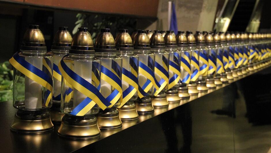 Fotografia przedstawia rząd identycznych zniczy ustawionych jeden obok drugiego na czarnej powierzchni. Każdy znicz jest ozdobiony przewiązaną wstążką w kolorach niebieskim i żółtym, co nawiązuje do barw Ukrainy. Znicze ustawione są w długiej, równiej linii, tworząc wizualnie efektowną kompozycję. Tło jest ciemne i nieostre, akcentując pierwszy plan.