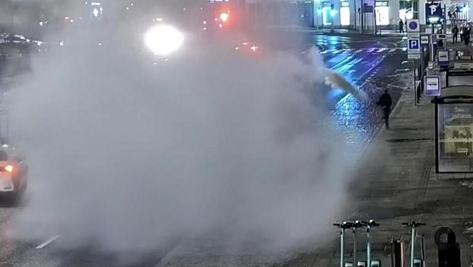 Na zdjęciu widać miejski chodnik oraz fragment ulicy nocą lub wczesnym wieczorem, oświetlone sztucznym oświetleniem. Znaczna część kadru po lewej stronie jest zasłonięta przez dużą chmurę gęstej mgły lub pary, która unosi się nad jezdnią. Źródło pary znajduje się z prawej strony – widać strumień skierowany w stronę ulicy.
Na prawym skraju obrazu stoi osoba na chodniku, odwrócona bokiem, w pobliżu miejsca, z którego wydobywa się para. Widać również wiatę przystankową oraz kilka stojących elektrycznych hulajnóg. Jezdnia jest mokra, co sugeruje deszcz lub świeżo zakończone opady. W tle widoczne są jasne światła pojazdów oraz budynki po drugiej stronie ulicy.