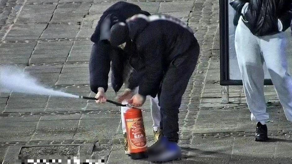 Na zdjęciu widać dwie osoby stojące na chodniku, pochylone nad gaśnicą ustawioną pionowo na ziemi. Jedna z osób trzyma w dłoniach wąż gaśnicy, z którego pod dużym ciśnieniem wydobywa się strumień białej substancji. Druga osoba trzyma korpus gaśnicy i manipuluje przy jej górnej części.
W tle, po prawej stronie kadru, widoczna jest trzecia osoba stojąca obok wiaty przystankowej. Chodnik jest wykonany z dużych płyt, a na krawędzi zdjęcia można dostrzec fragment drogi.
Scena odbywa się na zewnątrz, prawdopodobnie w przestrzeni miejskiej.