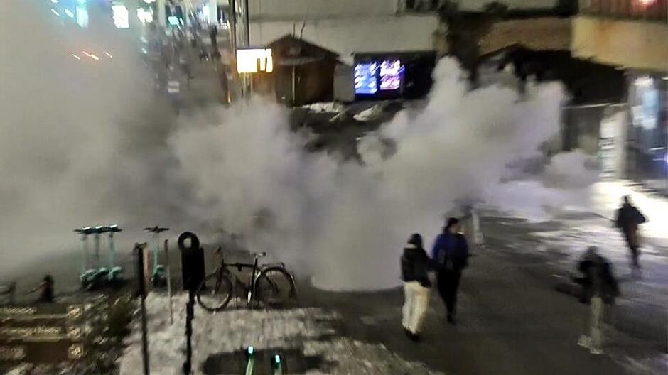 Zdjęcie przedstawia scenę miejską nocą lub wieczorem, oświetloną sztucznym światłem. Na środku kadru unosi się duża, gęsta chmura białego dymu lub pary, która zajmuje znaczną część przestrzeni i przysłania część tła.
Na pierwszym planie, po lewej stronie, widoczne są stojące elektryczne hulajnogi oraz zaparkowany rower. Na ziemi znajduje się również warstwa śniegu lub rozmokłego lodu.
Po prawej stronie obrazu widać kilka osób idących chodnikiem, częściowo oświetlonych i skierowanych w stronę od centrum zdjęcia. Tło obejmuje zabudowania oraz oświetlone tablice, jednak część szczegółów jest zakryta przez unoszącą się chmurę.
Cała scena wygląda na nagranie z monitoringu lub zdjęcie wykonane z wyższego punktu obserwacji.