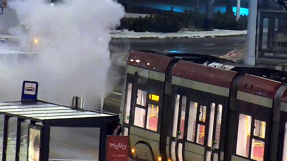 Zdjęcie przedstawia fragment miejskiego przystanku oraz przejeżdżający tramwaj. Kadrowanie obejmuje część wagonu z oświetlonymi wnętrzami oraz drzwiami. Po lewej stronie widoczna jest wiata przystankowa z tabliczką informującą o zatrzymujących się tu pojazdach komunikacji miejskiej.
Obok wiaty unosi się duża chmura białej pary lub dymu, która zasłania część ulicy i tła. Para wydobywa się z jednego, wyraźnie widocznego punktu przy krawędzi jezdni. W tle widać ośnieżone lub oblodzone fragmenty terenu oraz elementy infrastruktury drogowej.
Scena wygląda na ujęcie wykonane po zmroku, oświetlone sztucznym światłem, z możliwymi śladami zimowej pogody.