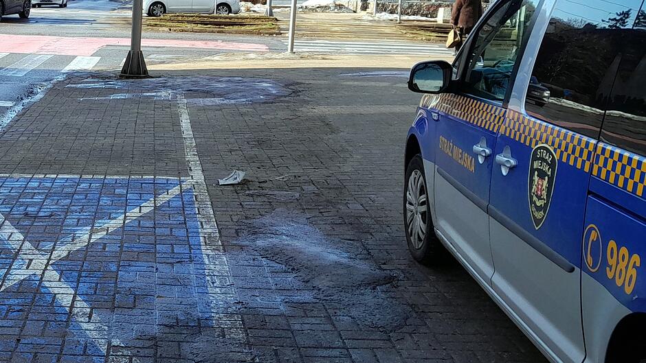 Na zdjęciu widać fragment chodnika oraz miejsce parkingowe oznaczone niebieską farbą. Na środku chodnika wystaje uszkodzony, metalowy element — wygląda jak pozostałość po odciętym słupku lub podstawie znaku. Obok stoi pojazd oznakowany jako Straż Miejska. W tle widać przejście dla pieszych, drogę oraz fragment mijających samochodów. Nawierzchnia jest miejscami zabrudzona i wilgotna.