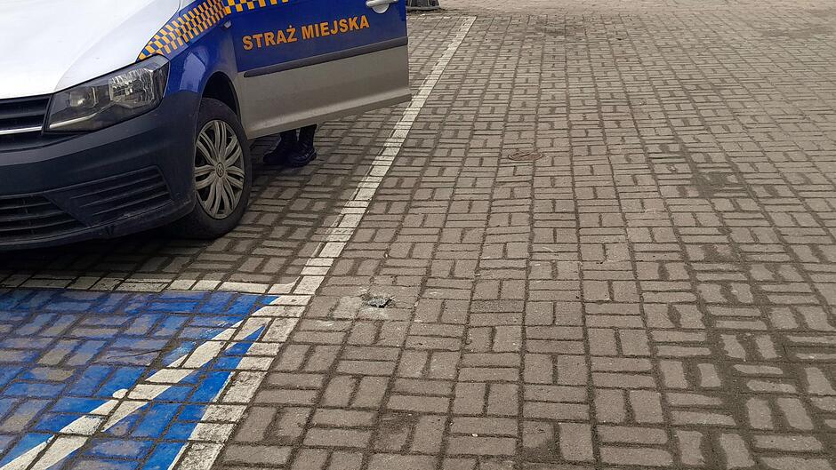Na zdjęciu widać fragment pojazdu oznakowanego jako Straż Miejska, zaparkowanego przy krawędzi miejsca postojowego. Drzwi pojazdu są otwarte, a na chodniku z kostki brukowej widoczny jest niewielki, uszkodzony element wystający z nawierzchni — wygląda to na pozostałość po odciętym słupku lub metalowej podstawie. Na pierwszym planie widać również fragment niebieskiego oznaczenia miejsca parkingowego. Cała scena znajduje się na utwardzonej powierzchni z kostki.