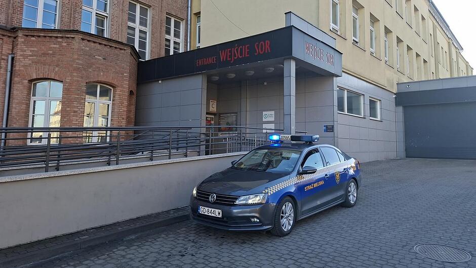 radiowóz straży miejskiej zaparkowany na podjeździe przed szpitalem na dachu ma włączone niebieskie światła w tle widać ceglany budynek i bramę z czerwonym napisem SOR