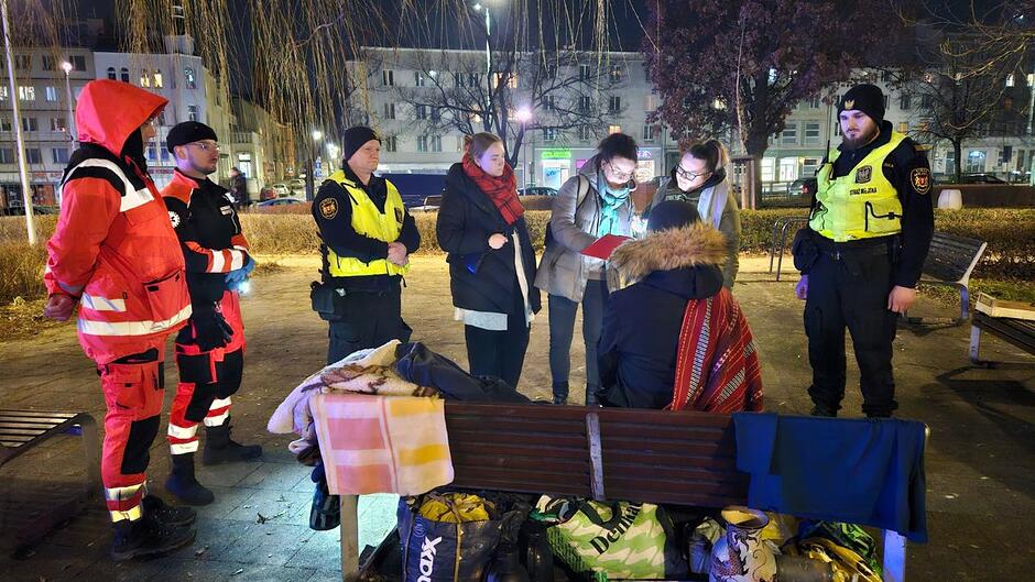 Strażnicy miejscy, ratownicy medyczni i wolontariusze stoją wokół osoby w kryzysie bezdomności siedzącej na ławce. Przeprowadzają z nią ankietę. 