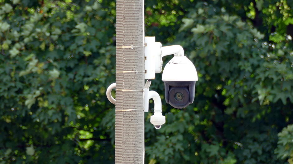 Kamera monitoringu miejskiego zamontowana na słupie, skierowana w stronę przestrzeni publicznej. W tle widoczne są zielone liście drzew.