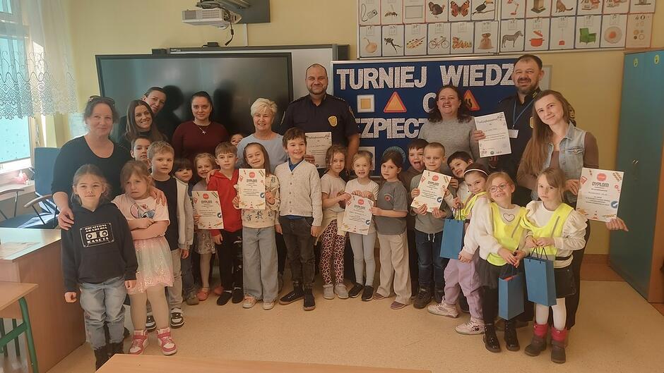 Przed czarną tablicą multimedialną stoi duża grupa dzieci wraz z kilkoma dorosłymi, prawdopodobnie opiekunami i przedstawicielami służb mundurowych. Na tablicy w tle widnieje napis: „TURNIEJ WIEDZ… BEZPIECZ…”, co może oznaczać turniej wiedzy o bezpieczeństwie. Wiele dzieci trzyma w rękach dyplomy i nagrody, a niektóre także kolorowe upominki. Dekoracje i ustawienie grupy wskazują na uroczyste zakończenie konkursu.