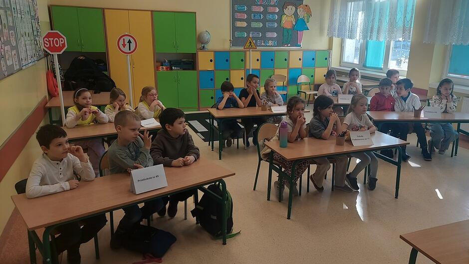 W sali lekcyjnej ustawione są trzy rzędy ławek, przy których siedzi grupa dzieci. Na stołach stoją kartoniki z nazwami drużyn, co sugeruje udział w konkursie lub zajęciach grupowych. W tle znajdują się kolorowe szafki w barwach zielonych, żółtych i pomarańczowych. Po lewej stronie widać tabliczkę „STOP” oraz znak nakazu kierunku, a na ścianie z prawej strony duże, słoneczne okna. Atmosfera wskazuje na edukacyjne wydarzenie związane z bezpieczeństwem lub ruchem drogowym.