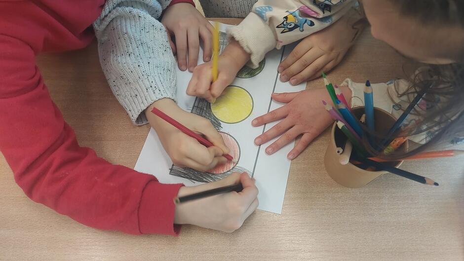 Z bliska pokazane są dłonie kilku dzieci, które kolorują rysunek sygnalizatora świetlnego. Na obrazku widoczne są trzy okręgi – czerwony, żółty i zielony. Dzieci używają kredek ołówkowych, a obok leży kubek pełen kredek. Zdjęcie przedstawia element zajęć plastycznych lub ćwiczenia dotyczącego zasad ruchu drogowego.