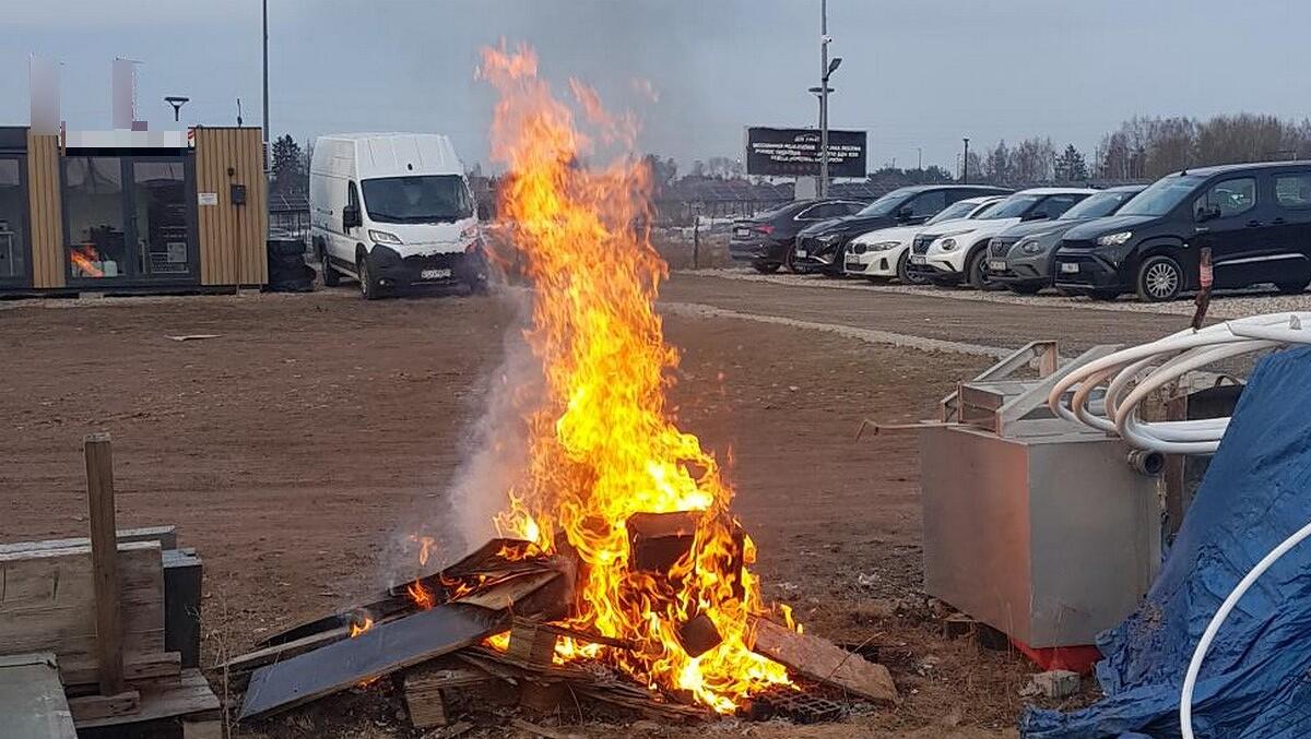 Na pierwszym planie widoczne jest duże, intensywnie płonące ognisko. Ogień ma wysokie, jasnopomarańczowe płomienie, unoszące się pionowo w górę. W palenisku znajdują się różne kawałki drewna oraz metalowe elementy – wyglądają na odpady budowlane lub rozbiórkowe.
Ognisko znajduje się na nieutwardzonym, ziemistym terenie, przypominającym plac budowy lub zaplecze techniczne. W pobliżu widać: po lewej stronie: stosy desek i elementów konstrukcyjnych,
po prawej stronie: metalowa skrzynia z podłączonymi białymi przewodami i przykryta niebieską plandeką,
w tle: zaparkowane samochody osobowe ustawione w rzędzie,
jeszcze dalej: jasny samochód dostawczy stojący obok małego kontenera lub budynku modułowego, na którym widać okno i drzwi.

Niebo jest szare i zachmurzone, co sugeruje pochmurny, chłodny dzień. Całość sprawia wrażenie miejsca pracy lub placu budowy, gdzie spalane są materiały odpadowe.