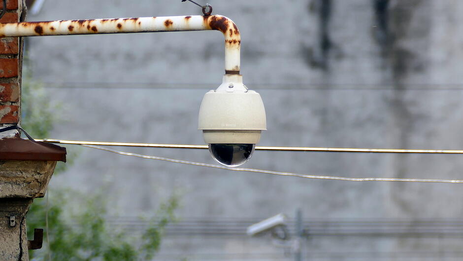 Na zdjęciu widoczna jest zamontowana na metalowym, częściowo zardzewiałym wysięgniku kamera monitoringu. Urządzenie ma kopułkową obudowę i skierowane jest w dół. W tle znajduje się rozmyty, szary mur oraz przewody biegnące poziomo przez kadr. W dolnej części widoczna jest także kolejna kamera umieszczona w oddali.