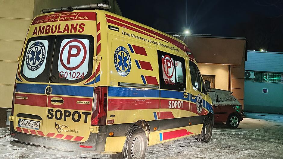 Na zdjęciu widoczny jest ambulans stojący na ośnieżonej nawierzchni, w pobliżu budynku o jasnych ścianach. Pojazd jest żółto‑czerwony, oznakowany jako ambulans ratownictwa medycznego z Sopotu. Na tylnej części karetki umieszczone są symbole medyczne, oznaczenia pojazdu, litera „P” w czerwonym okręgu oraz informacja o sfinansowaniu zakupu z budżetu obywatelskiego. Obok karetki stoi czerwony samochód osobowy. Scena ma charakter nocny — oświetlenie pochodzi z lamp zewnętrznych przy budynku.