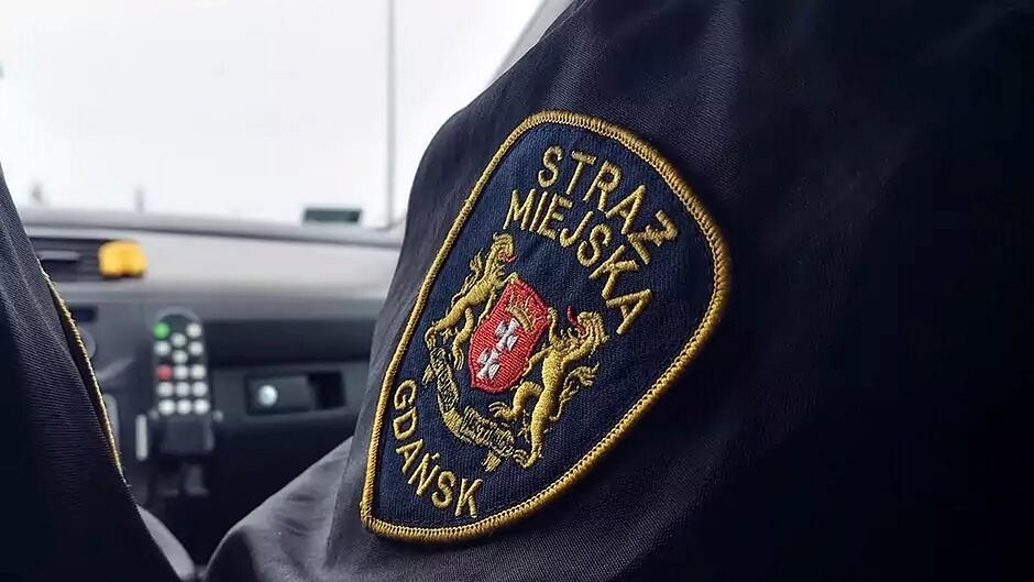 Na zdjęciu widać fragment ciemnego umundurowania z naszytym emblematem Straży Miejskiej w Gdańsku. Herb na naszywce przedstawia czerwone pole z białym motywem oraz dwie złote sylwetki lwów po bokach. Emblemat ma kształt tarczy z żółtym obramowaniem i wyraźnymi napisami: „STRAŻ MIEJSKA” u góry oraz „GDAŃSK” na dole. W tle znajduje się wnętrze pojazdu służbowego z widoczną deską rozdzielczą i zamontowanym radiotelefonem.