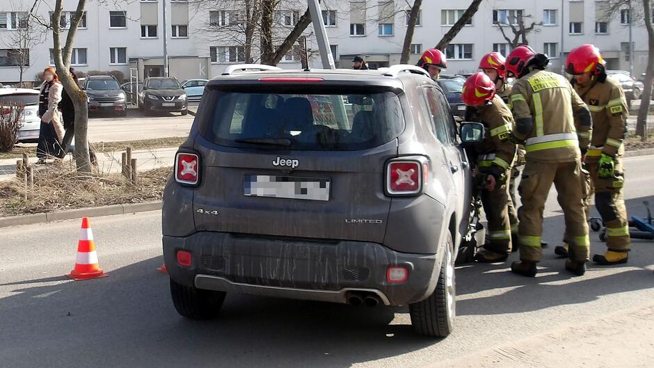 Na zdjęciu widać pojazd marki Jeep zatrzymany na jezdni w pobliżu krawężnika. Obok samochodu znajduje się grupa osób ubranych w specjalistyczne ubrania ochronne i kaski, typowe dla służb ratowniczych. Pochylają się nad osobą lub sprzętem znajdującym się na ziemi, wykonując czynności pomocowe. Na drodze ustawiono pomarańczowo‑białą pachołek drogowy, zabezpieczający miejsce zdarzenia. W tle widoczne są zaparkowane samochody, chodnik oraz budynki mieszkalne.