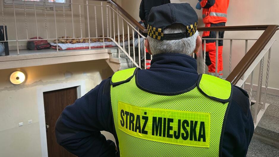Na zdjęciu widoczna jest postać w umundurowaniu Straży Miejskiej stojąca na klatce schodowej. Osoba ma na sobie jaskrawożółty odblaskowy bezrękawnik z dużym napisem „STRAŻ MIEJSKA” na plecach oraz czapkę z charakterystyczną szachownicą. W tle, na wyższym poziomie klatki schodowej, znajdują się kolejne osoby ubrane w odzież ochronną w kolorze pomarańczowym. Obok nich prowadzone są czynności służbowe. Przy balustradzie widoczne jest miejsce z położonymi kocami i materacem. Ściany i schody wskazują na wnętrze wielopiętrowego budynku.