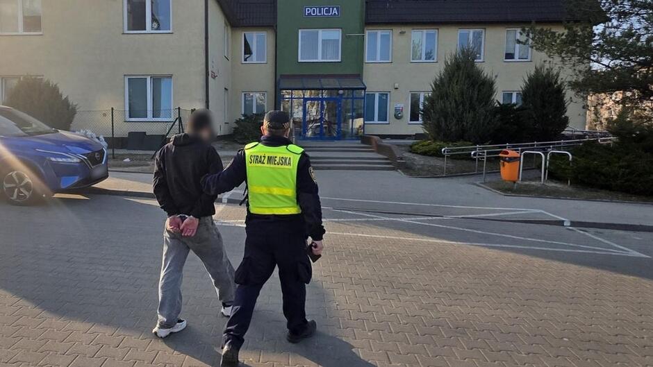 zdjęcie przedstawia scenę na zewnątrz budynku oznaczonego szyldem „POLICJA”, znajdującego się na wprost w tle. Budynek jest dwukondygnacyjny, w jasnym kolorze, z zielonym elementem fasady wokół wejścia. Wejście znajduje się pośrodku i prowadzą do niego niewysokie schody oraz podjazd. Przed budynkiem widoczne są krzewy i elementy zagospodarowania terenu, m.in. kosz na śmieci w kolorze pomarańczowym.
Na pierwszym planie, z lewej strony, znajduje się pojazd w kolorze niebieskim. Obok niego, na środku zdjęcia, widoczne są dwie osoby: jedna z nich prowadzi drugą w kierunku budynku. Osoba po prawej stronie ma na sobie odblaskową kamizelkę z napisem „STRAŻ MIEJSKA” na plecach oraz ciemny mundur. Druga osoba ma związane ręce z tyłu i twarz rozmytą (nieczytelną), oraz ubranie w ciemną górę i jasne spodnie. Obydwie osoby idą po brukowanej nawierzchni w kierunku wejścia posterunku.