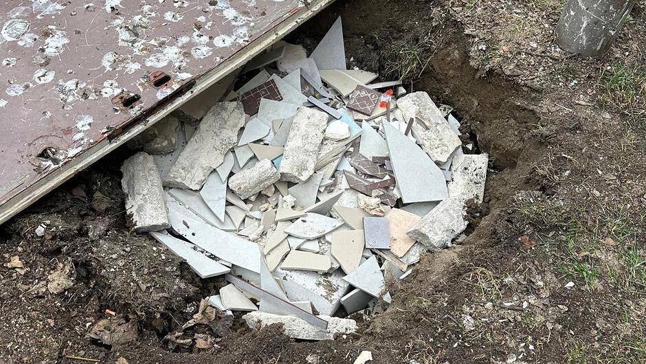 Na zdjęciu widoczny jest spory wykop w ziemi znajdujący się na terenie porośniętym trawą. Wewnątrz wykopu znajdują się liczne odpady poremontowe, takie jak potłuczone płytki ceramiczne, fragmenty betonu, kawałki zaprawy oraz inne elementy gruzu o różnych kształtach i rozmiarach. Nad lewą częścią wykopu opiera się duży, prostokątny element przypominający uszkodzone drzwi lub płytę, z licznymi odpryskami i śladami zużycia na powierzchni. Krawędzie ziemi wokół dołu są odsłonięte i nieregularne, co wskazuje na niedawne naruszenie gruntu. Po prawej stronie widoczny jest fragment stalowego słupa lub rury.