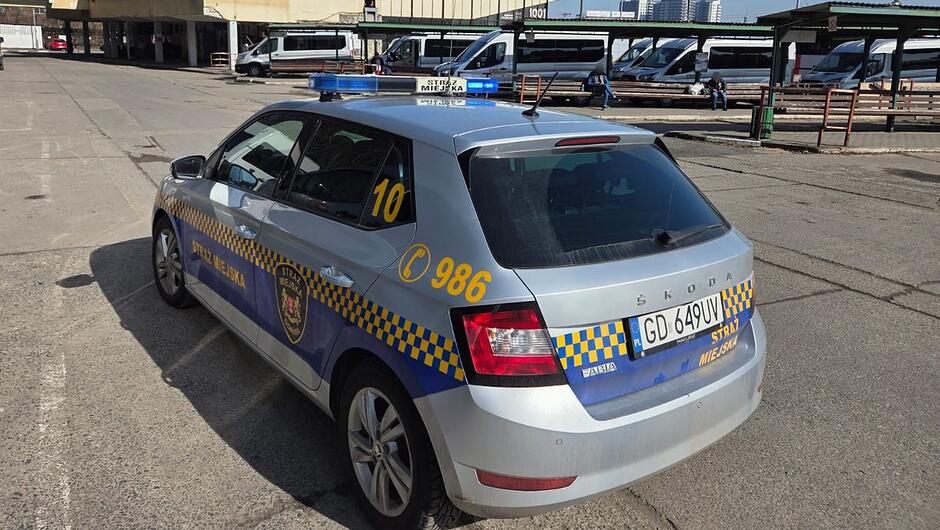 Na zdjęciu widać samochód Straży Miejskiej zaparkowany na terenie przypominającym dworzec autobusowy lub miejskie centrum przesiadkowe. Pojazd to srebrna Škoda Fabia z charakterystycznymi niebiesko‑żółtymi oznaczeniami, numerem bocznym 10 i numerem alarmowym 986. Na dachu znajduje się belka świetlna.
W tle stoją minibusy i busy, a w okolicy widać kilka osób – część siedzi na ławkach, inni przechodzą obok. Otoczenie sugeruje typową miejską przestrzeń publiczną, gdzie obecność straży miejskiej ma podkreślać nadzór nad porządkiem i bezpieczeństwem.