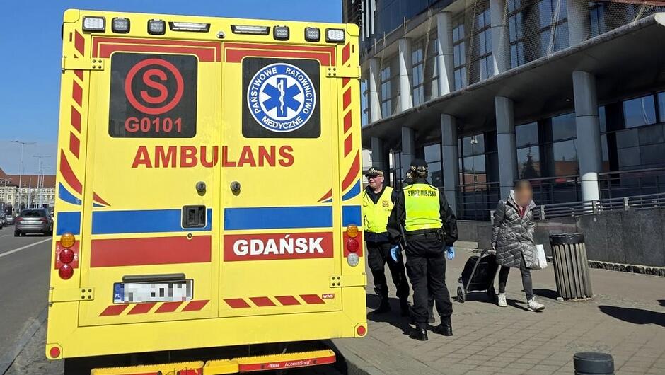 Na zdjęciu widać grupę funkcjonariuszy straży miejskiej oraz pracowników ratownictwa medycznego stojących na chodniku przy budynku z dużymi szybami. W centrum znajduje się osoba siedząca na ziemi, a ratownicy i strażnicy są pochylani w jej kierunku, udzielając pomocy. W tle widoczne są barierki, elewacja budynku oraz przechodnie znajdujący się po drugiej stronie ulicy.