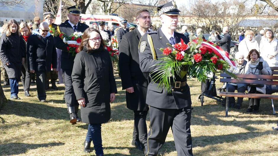 Strażnik miejski niesie wiązankę kwiatów, za nim idzie dwuosobowa delegacja Miasta Gdańska: radna Kamila Błaszczak i zastępca prezydenta Gdańska Piotr Borawski. W tle zgromadzona publiczność. 