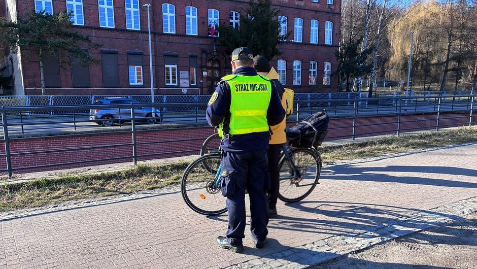 Na pierwszym planie widać dwie osoby stojące na utwardzonej ścieżce pieszo‑rowerowej. Jedna z tych osób ma na sobie odblaskową kamizelkę z napisem „STRAŻ MIEJSKA” na plecach oraz ciemne spodnie i czapkę. Druga osoba stoi obok roweru, który ma zamontowany bagażnik z tyłu.
Obie postacie zwrócone są tyłem do obiektywu. Osoba w kamizelce stoi najbliżej aparatu, a obok niej znajduje się rower oraz druga osoba ubrana w kurtkę w jasnym kolorze.
W tle widoczny jest czerwony, kilkupiętrowy budynek z cegły, z dużymi jasnymi oknami. Przed budynkiem biegnie uliczka, po której jedzie samochód. Wzdłuż chodnika i kanału (lub obniżonego terenu oddzielonego murkiem i barierką) przebiega metalowa balustrada. Po prawej stronie w tle znajdują się drzewa o bezlistnych gałęziach, co sugeruje porę późnojesienną lub zimową. Oświetlenie jest słoneczne, a cienie są wyraźne.
