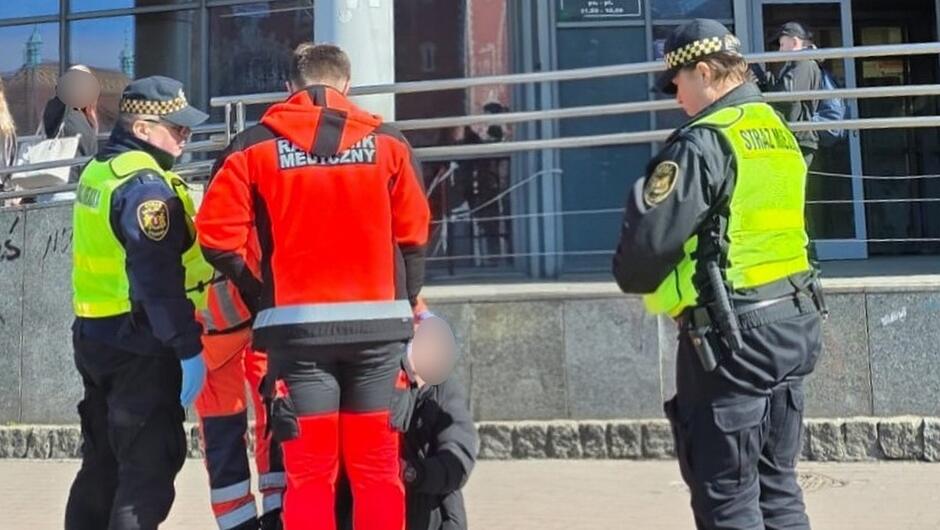 Na zdjęciu widać grupę funkcjonariuszy straży miejskiej oraz pracowników ratownictwa medycznego stojących na chodniku przy budynku z dużymi szybami. W centrum znajduje się osoba siedząca na ziemi, a ratownicy i strażnicy są pochylani w jej kierunku, udzielając pomocy. W tle widoczne są barierki, elewacja budynku oraz przechodnie znajdujący się po drugiej stronie ulicy.