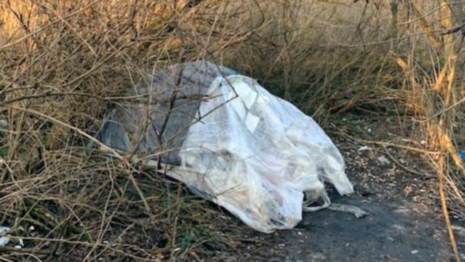 Na zdjęciu widać skupisko suchych, gęstych gałęzi i zarośli. W centrum znajduje się duży stos odpadów owiniętych w przezroczystą folię, leżący bezpośrednio na ziemi. Folia jest częściowo zgnieciona i nieprzezroczysta w niektórych miejscach, a spod niej wystają fragmenty różnych materiałów. Dookoła widać ścieżkę ziemną oraz pojedyncze drobne śmieci. Tło stanowią wysokie, suche trawy i krzewy.