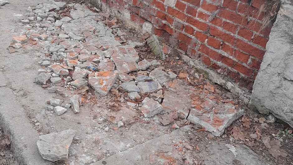 Na zdjęciu widać fragment zniszczonego chodnika przylegającego do ceglanego muru. Powierzchnia chodnika jest mocno uszkodzona — liczne płyty i elementy betonowe są popękane, pokruszone i porozrzucane. Wśród gruzu widoczne są kawałki cegieł oraz fragmenty pokryte pomarańczowym pyłem. Między szczelinami leżą suche liście i drobne zanieczyszczenia. Ceglana ściana w tle ma miejscami ubytki i nierówności, a obok niej widoczny jest fragment betonowego elementu konstrukcyjnego.