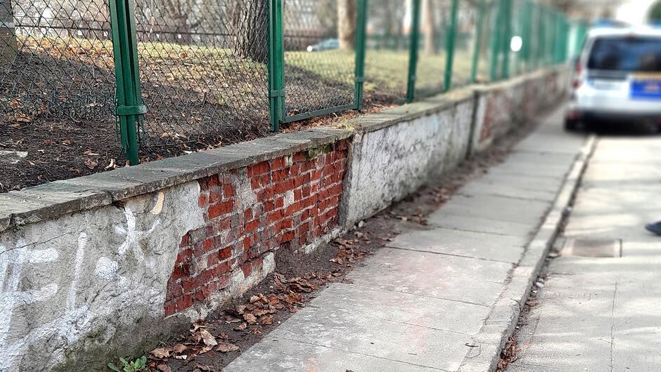 Na zdjęciu widoczny jest fragment chodnika biegnący wzdłuż niskiego muru z cegły i betonu. Ściana ma liczne ubytki — miejscami odsłonięte są czerwone cegły, a w innych miejscach widać kruszącą się, jasną warstwę tynku. Przy murze leży warstwa suchych liści. Po lewej stronie znajdują się zielone metalowe słupki ogrodzenia z umocowaną siatką. Za ogrodzeniem widoczny jest teren zielony z drzewami. Po prawej stronie kadru widać zaparkowany samochód częściowo wychodzący poza obraz.