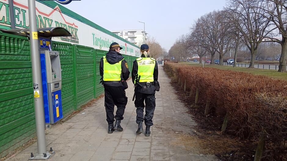Na zdjęciu widać dwóch umundurowanych funkcjonariuszy straży miejskiej idących chodnikiem wzdłuż zielonego ogrodzenia targowiska. Funkcjonariusze są sfotografowani od tyłu i mają na sobie odblaskowe kamizelki z napisem „STRAŻ MIEJSKA”. Po lewej stronie kadru znajduje się ściana targowiska w zielonym kolorze oraz wolnostojący parkometr. Po prawej stronie widoczny jest rząd niskich, przyciętych krzewów, za którymi rozciąga się teren zielony z drzewami. W oddali, po prawej stronie, stoi zaparkowany samochód. Chodnik jest szeroki i biegnie prosto w kierunku dalszej części osiedla i zabudowań widocznych w tle.