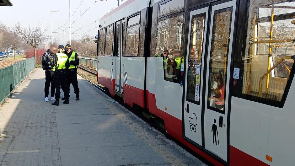 Na zdjęciu widoczna jest sytuacja na przystanku tramwajowym. Przy krawędzi peronu stoi tramwaj w barwach białej i czerwonej, z otwartymi drzwiami. Obok tramwaju znajduje się grupa osób: dwie umundurowane osoby w odblaskowych kamizelkach oraz jedna osoba w ciemnym ubraniu. Osoby stoją na chodniku wykonanym z betonowych płyt. W tle po lewej stronie widać zielone ogrodzenie oraz rząd krzewów. Dalej, wzdłuż torowiska, rozciąga się linia trakcyjna i widoczne są drzewa bez liści, a po lewej stronie kadru – fragment jezdni z poruszającymi się pojazdami. Scena przedstawia kontrolę lub interwencję służb przy zatrzymanym tramwaju.