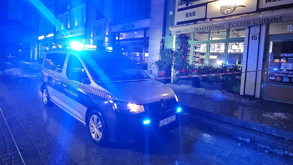 Na zdjęciu widać oznakowany pojazd Straży Miejskiej stojący na ulicy w porze nocnej. Auto ma włączone niebieskie światła sygnalizacyjne, które oświetlają okolice. Po prawej stronie kadru znajduje się lokal gastronomiczny z widoczną witryną i oświetleniem wewnątrz. Przed lokalem ustawione jest czerwono-białe taśmowe wygrodzenie, sugerujące zabezpieczenie terenu. Na chodniku i przy krawężniku widać resztki śniegu. Tło stanowią kamienice i miejskie oświetlenie.