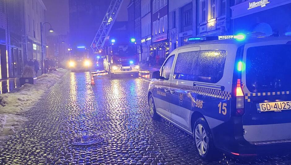 Na zdjęciu widać nocną scenę na brukowanej ulicy, na której stoją pojazdy służb. Po prawej stronie znajduje się oznakowany radiowóz Straży Miejskiej z włączonymi niebieskimi światłami sygnalizacyjnymi. W dalszej części ulicy, na wprost, widoczne są dwa wozy strażackie również z aktywnym oświetleniem błyskowym. Jeden z nich ma wysuniętą drabinę skierowaną ku górze. Ulica jest mokra, a odbicia świateł tworzą jasne refleksy na nawierzchni. Po bokach kadru widać kamienice, sklepy oraz fragmenty pozostawionego śniegu. Scena przedstawia działania służb w centrum miasta.