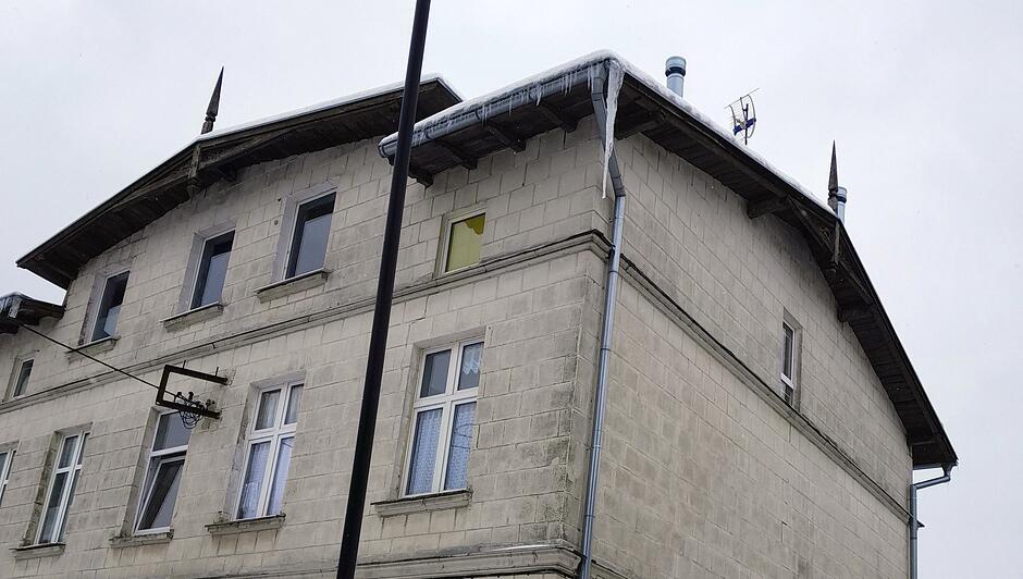 Na zdjęciu widać narożną część dwupiętrowego budynku wykonanego z jasnych, prostokątnych bloczków. Wzdłuż krawędzi dachu zwisa długi, masywny sopel lodu, a pod nim widoczna jest rynna. Na dachu znajdują się także elementy instalacji, w tym niewielka antena. Okna budynku są różnej wielkości; jedno z nich, na poddaszu, ma żółte zasłonięcie od środka. Na pierwszym planie stoi wysoka, czarna latarnia uliczna. Niebo jest szare i pochmurne, co sugeruje zimową aurę.