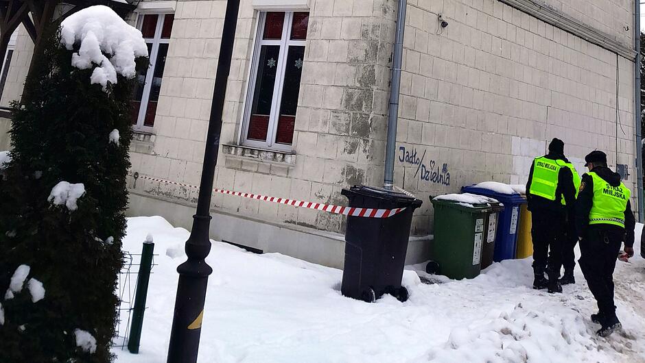 Na zdjęciu widać fragment budynku o jasnej elewacji oraz zasypany śniegiem teren wokół niego. Przy ścianie stoją trzy pojemniki na odpady w różnych kolorach: czarny, zielony i niebieski. Wzdłuż budynku rozciągnięta jest biało-czerwona taśma ostrzegawcza, zamocowana między pojemnikiem a elementem elewacji. Po prawej stronie kadru idą trzej umundurowani funkcjonariusze straży miejskiej w odblaskowych kamizelkach. Po lewej stronie widoczny jest ośnieżony krzew oraz latarnia uliczna. Na ścianie budynku znajduje się niewielki napis wykonany sprayem. Śnieg na ziemi jest udeptany, tworząc wąską ścieżkę.