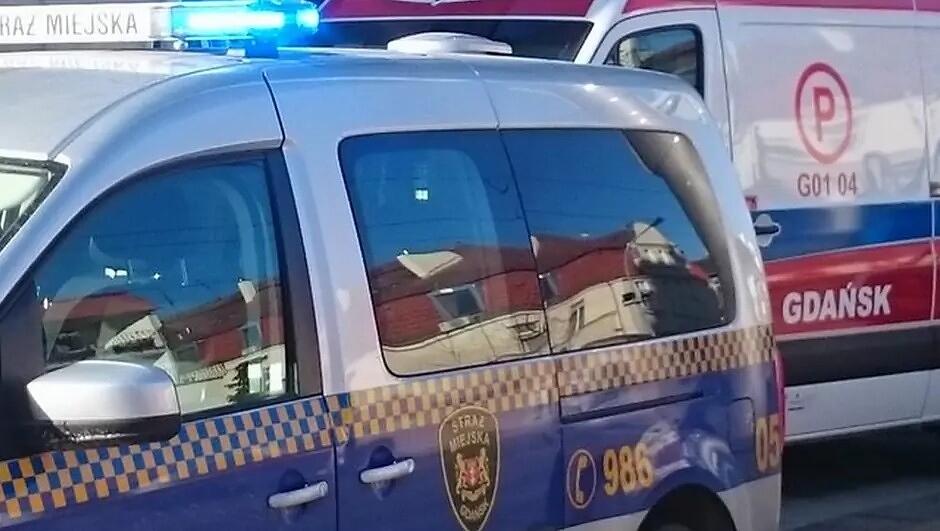 Na zdjęciu widoczny jest radiowóz straży miejskiej z włączonymi niebieskimi światłami sygnalizacyjnymi. Pojazd stoi obok karetki pogotowia ratunkowego oznaczonej napisem „GDAŃSK” i symbolem „P”. W odbiciach szyb radiowozu widać fragmenty pobliskich budynków. Oba pojazdy znajdują się bardzo blisko siebie, co sugeruje prowadzenie wspólnych działań lub interwencji. Scena ma miejsce w świetle dziennym.