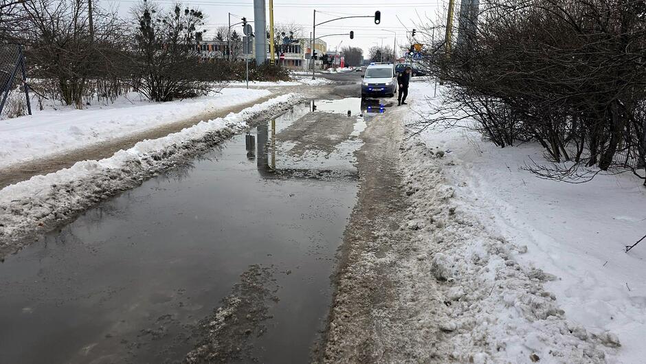 Na zdjęciu widać ośnieżoną, lokalną drogę, na której utworzyło się duże rozlewisko wodne. Woda zajmuje większość jezdni, tworząc szeroką, głęboką kałużę. Po prawej stronie drogi znajduje się wąski, udeptany pas śniegu, którym poruszają się dwie osoby stojące obok oznakowanego radiowozu straży miejskiej. Pojazd ma włączone niebieskie światła sygnałowe, odbijające się w powierzchni wody. W tle widoczne są sygnalizatory świetlne, słupy, niskie zabudowania oraz zarośla po obu stronach drogi. Scena przedstawia zimowe warunki i utrudnienia wynikające z roztopów.