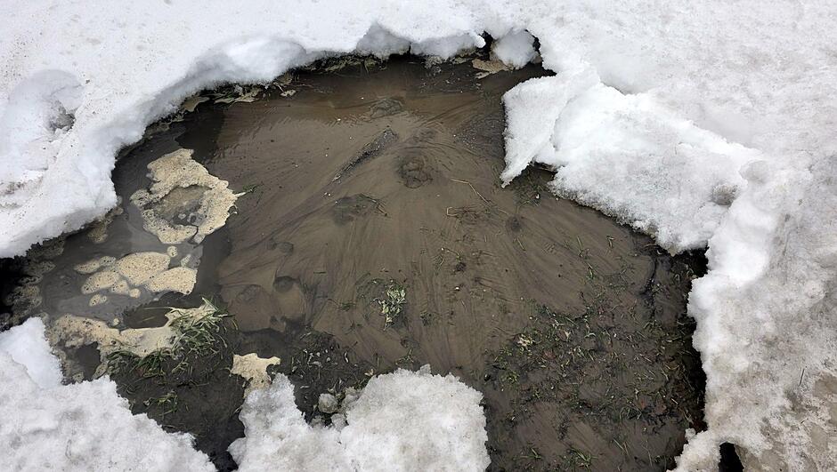 Na zdjęciu widać dużą kałużę brudnej, brunatnej wody powstałą w zagłębieniu terenu. Kałuża otoczona jest topniejącym śniegiem, którego krawędzie są nieregularne i podmyte wodą. W kilku miejscach spod śniegu wystają fragmenty ziemi oraz źdźbła trawy. Na powierzchni wody widoczne są osady ziemi i smugi tworzące charakterystyczne wzory. Scena przedstawia efekt roztopów i nagromadzenie błotnistej wody w obniżeniu terenu.