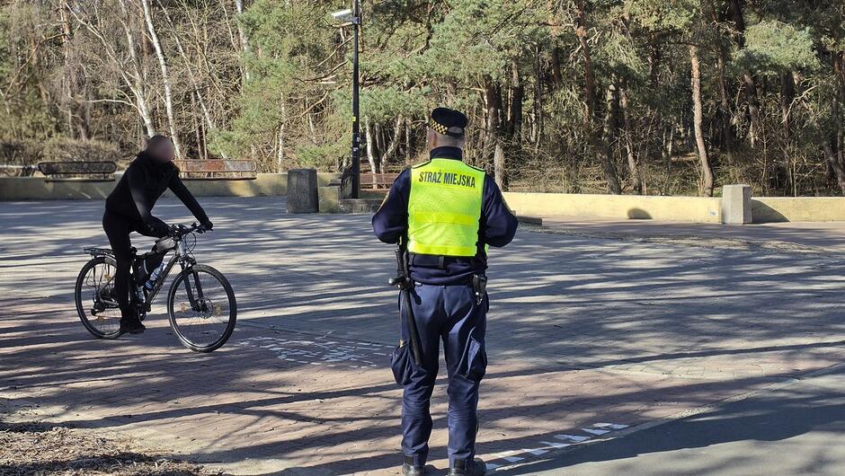 Na pierwszym zdjęciu widać funkcjonariusza Straży Miejskiej stojącego tyłem do obiektywu. Ma na sobie żółtą kamizelkę odblaskową z napisem „STRAŻ MIEJSKA”. Funkcjonariusz znajduje się na otwartej przestrzeni przy lesie lub parku. Przed nim jedzie rowerzystka ubrana na czarno, patrząca w stronę strażnika. W tle widać drzewa oraz elementy infrastruktury parkowej.